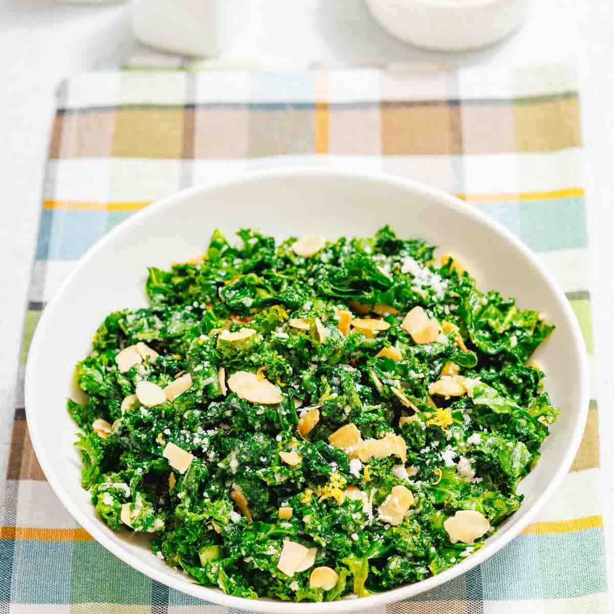 A white bowl filled with kale salad topped with sliced almonds and grated cheese, placed on a plaid cloth.