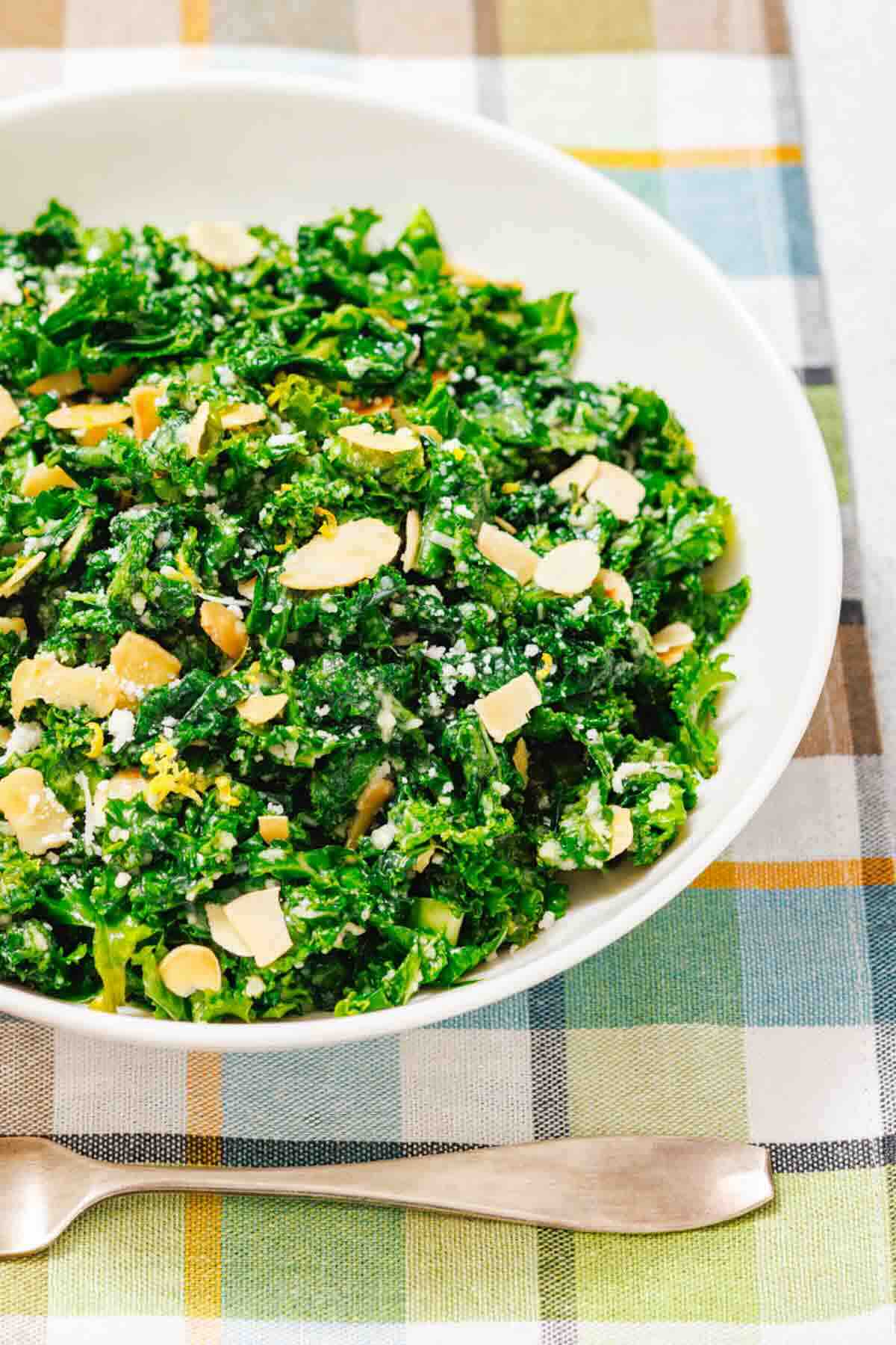 A white bowl of kale salad topped with sliced almonds and grated cheese sits on a plaid tablecloth beside a fork.