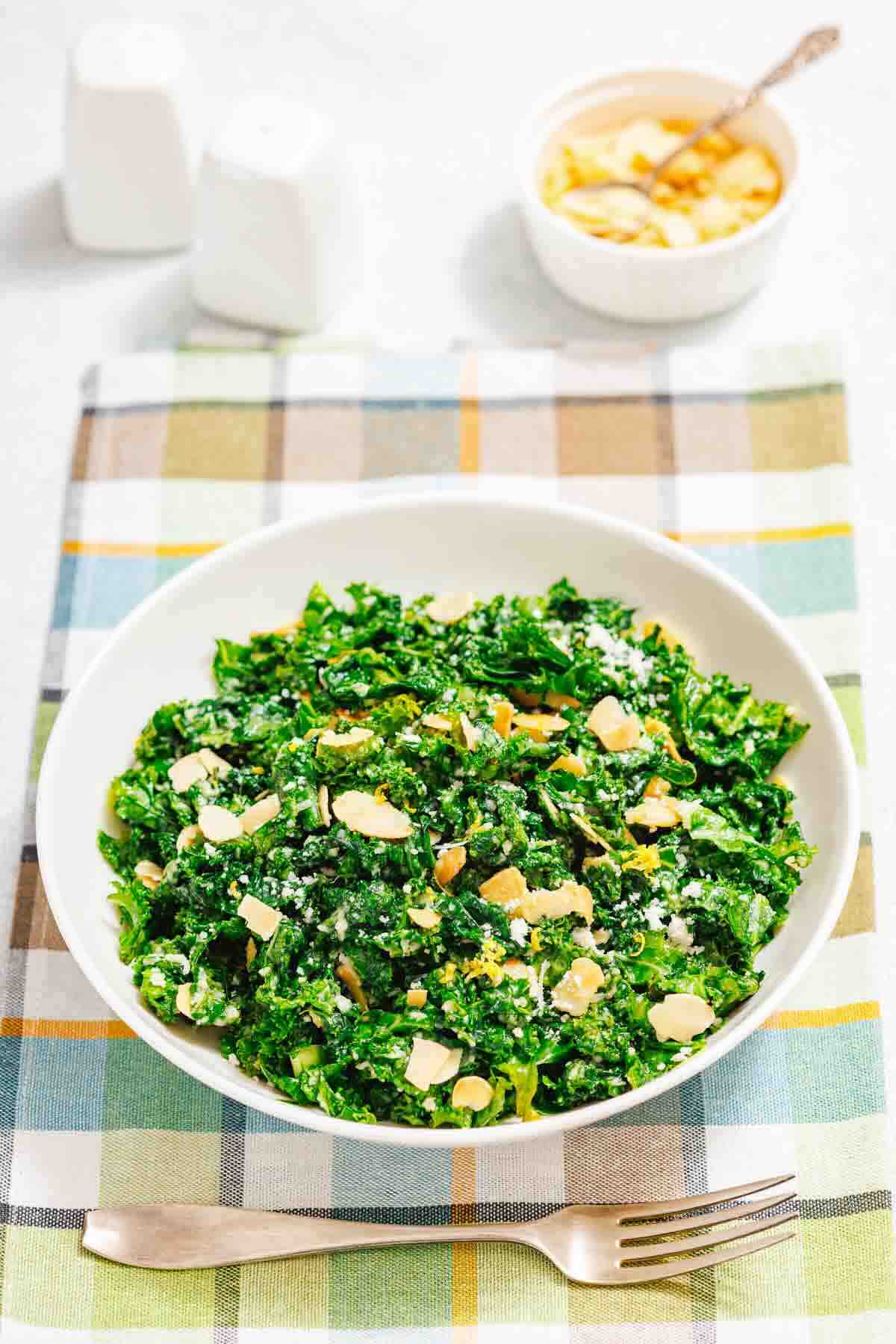 A white bowl of kale salad topped with sliced almonds is placed on a plaid cloth with a fork and spoon beside it. A small bowl of almonds is in the background.