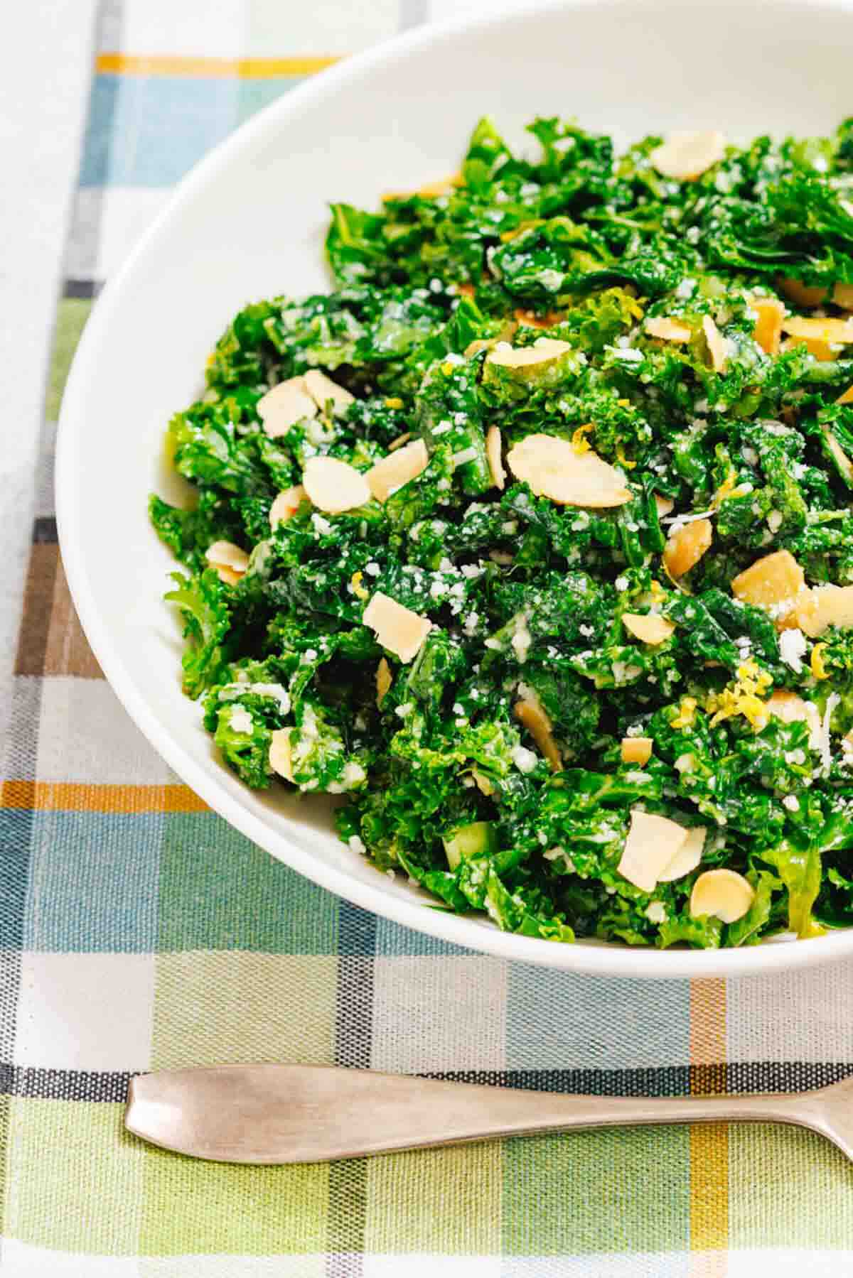 A white bowl filled with kale salad topped with sliced almonds and grated cheese, placed on a plaid tablecloth next to a silver utensil.