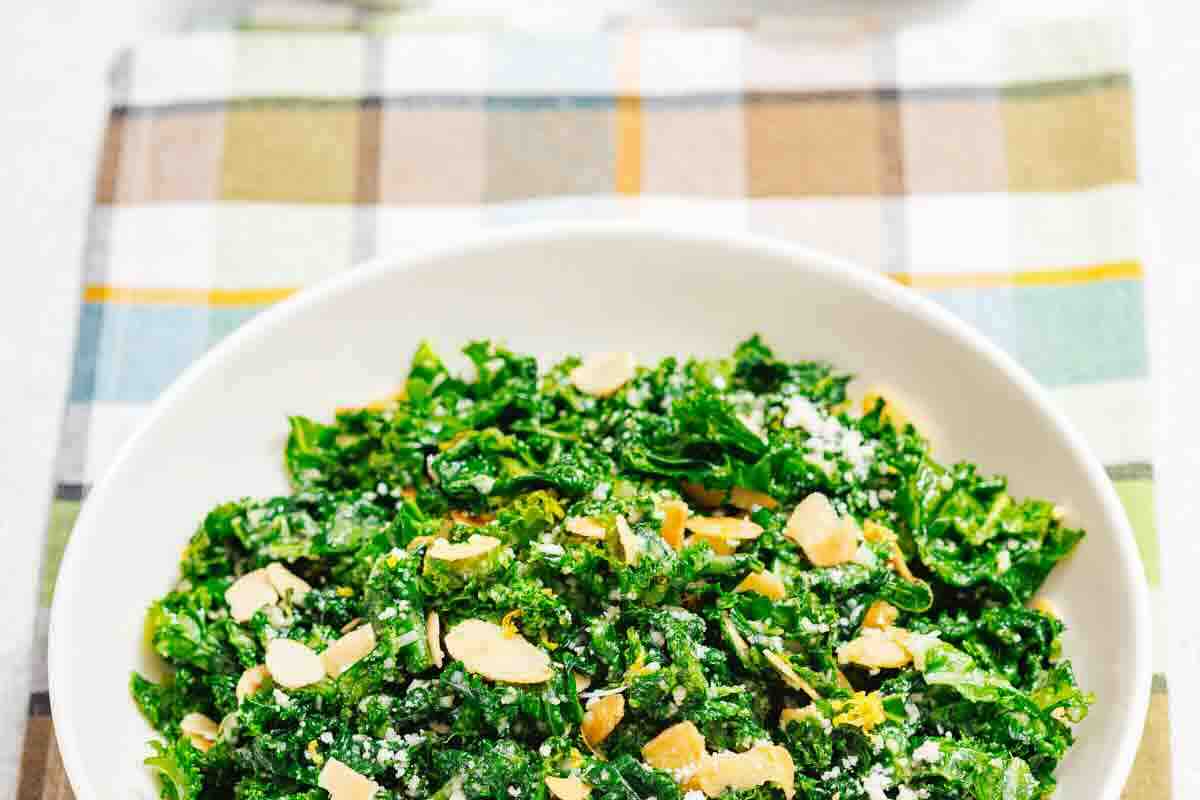 A white bowl filled with kale salad topped with sliced almonds and grated cheese, placed on a plaid cloth.