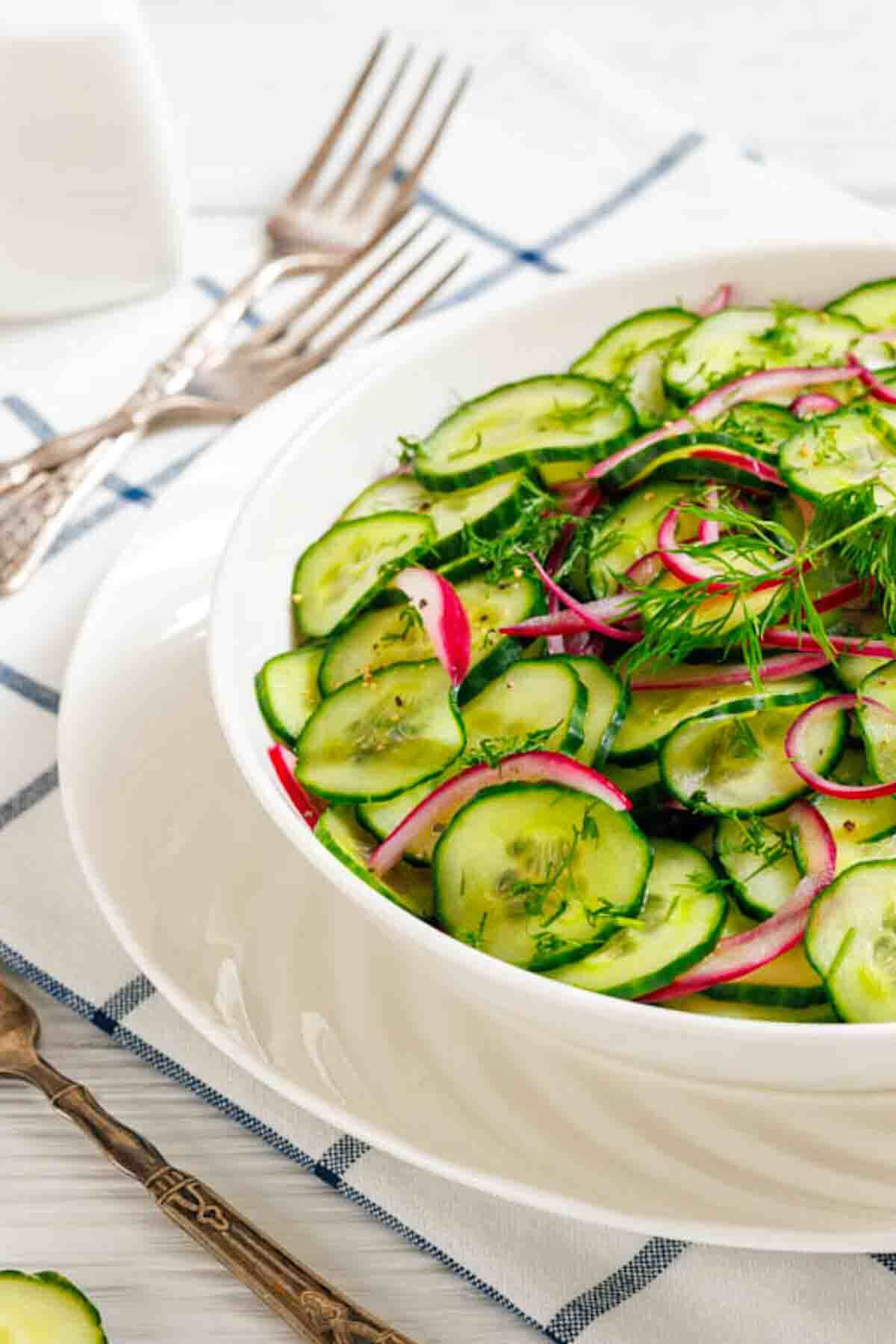 A white bowl filled with cucumber salad, red onions, and dill sits on a white plate with metal forks beside it on a checkered napkin.