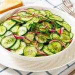 A white bowl filled with cucumber salad, garnished with dill and thin slices of red onion, on a checkered tablecloth with cutlery and toast in the background.