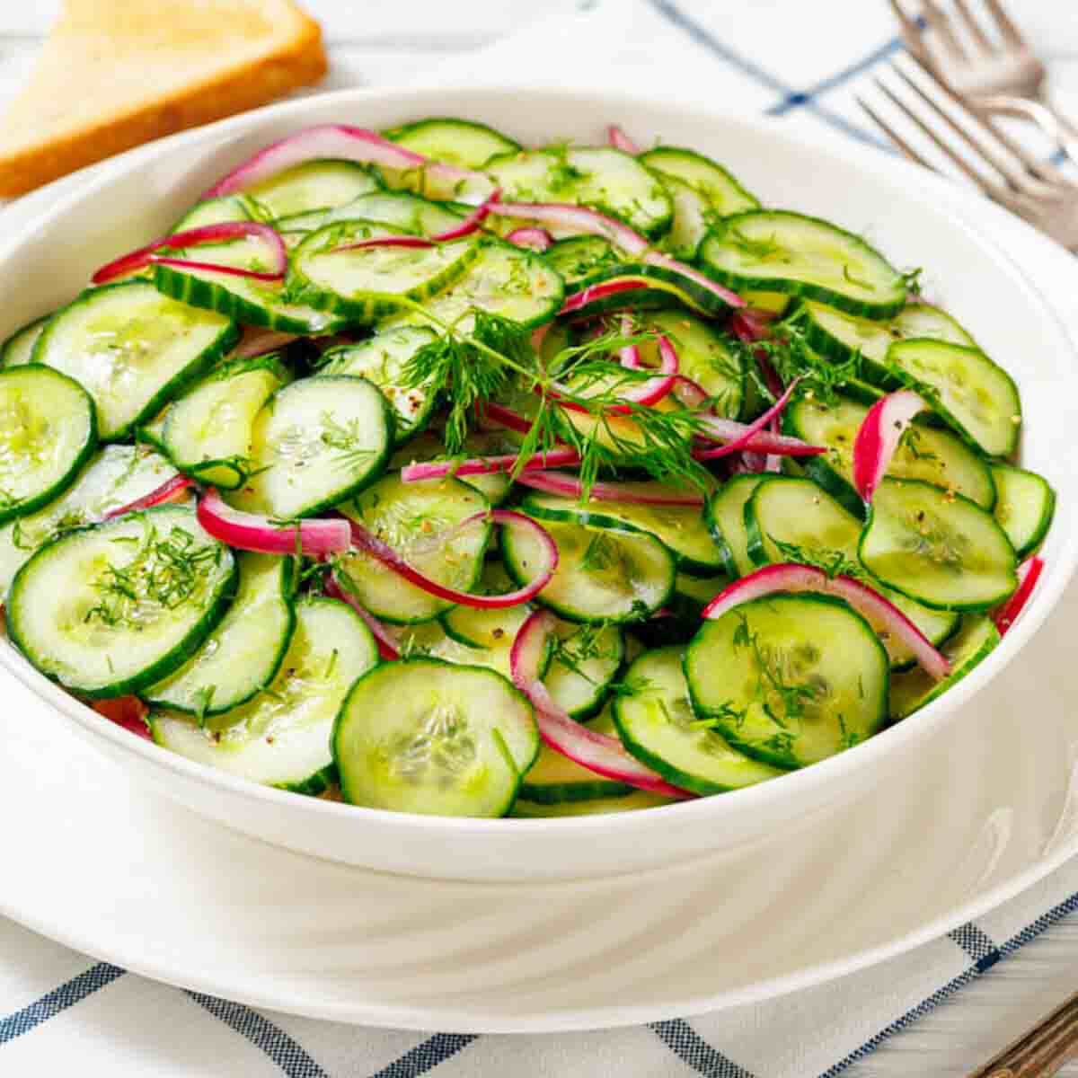 A white bowl filled with cucumber salad, garnished with dill and thin slices of red onion, on a checkered tablecloth with cutlery and toast in the background.
