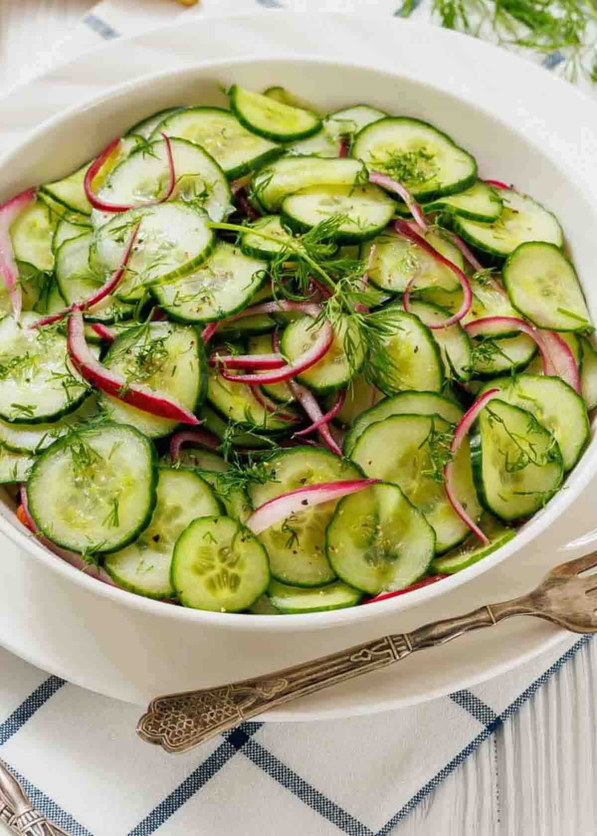 A bowl of cucumber salad with thinly sliced cucumbers, red onions, and fresh dill on a white plate, set on a checkered napkin with silverware nearby.