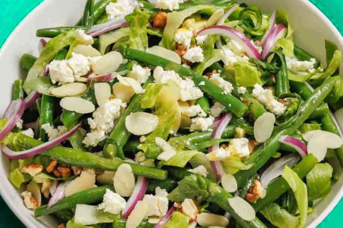 A bowl of green bean salad with sliced almonds, red onion, lettuce, walnuts, and crumbled feta cheese.
