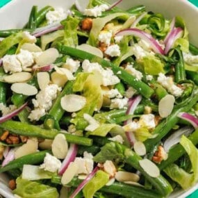 A salad with green beans, lettuce, sliced almonds, red onion, crumbled cheese, and walnuts in a white bowl.