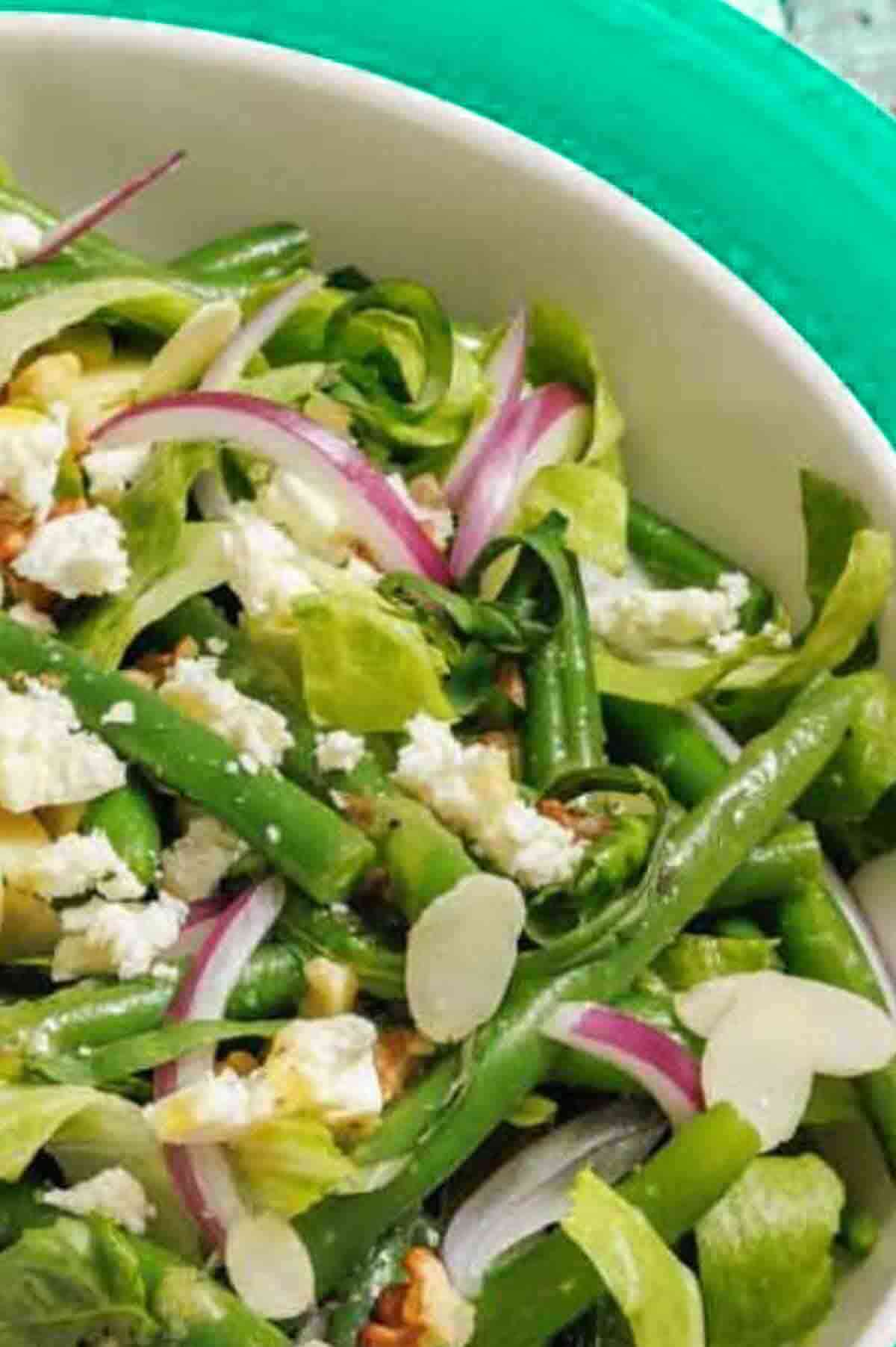 A salad with green beans, lettuce, sliced red onions, feta cheese, almond slivers, and walnuts in a white bowl.