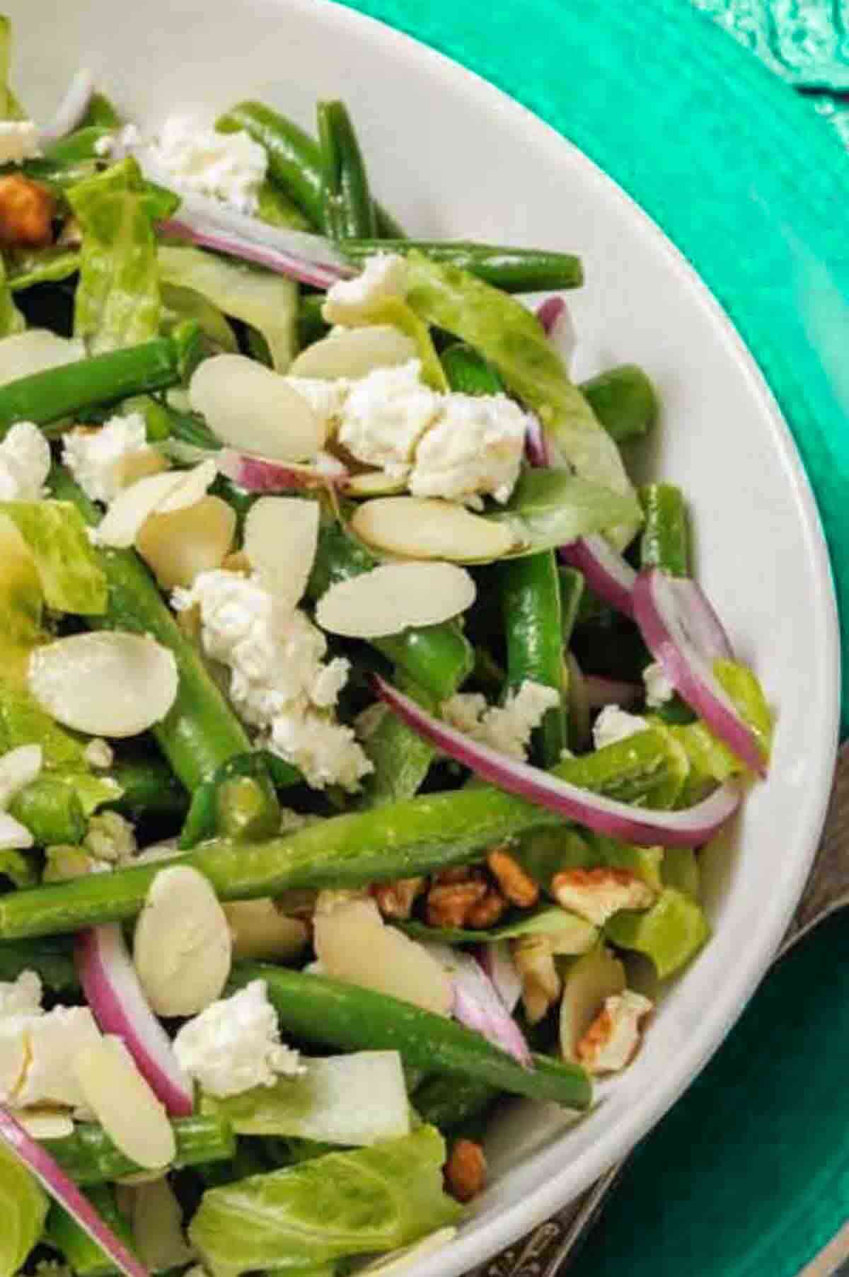 A bowl of salad with green beans, lettuce, sliced almonds, red onion, crumbled cheese, and walnuts on a turquoise background.