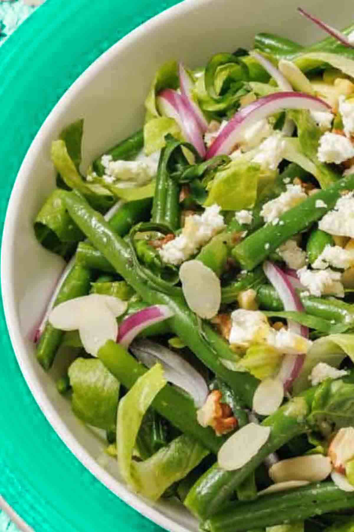 A bowl of green bean salad with sliced red onions, lettuce, crumbled cheese, almond slices, and walnut pieces on a green plate.