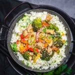 A bowl of cauliflower rice topped with stir-fried vegetables, sesame seeds, and pieces of tofu or chicken, garnished with chopped herbs.