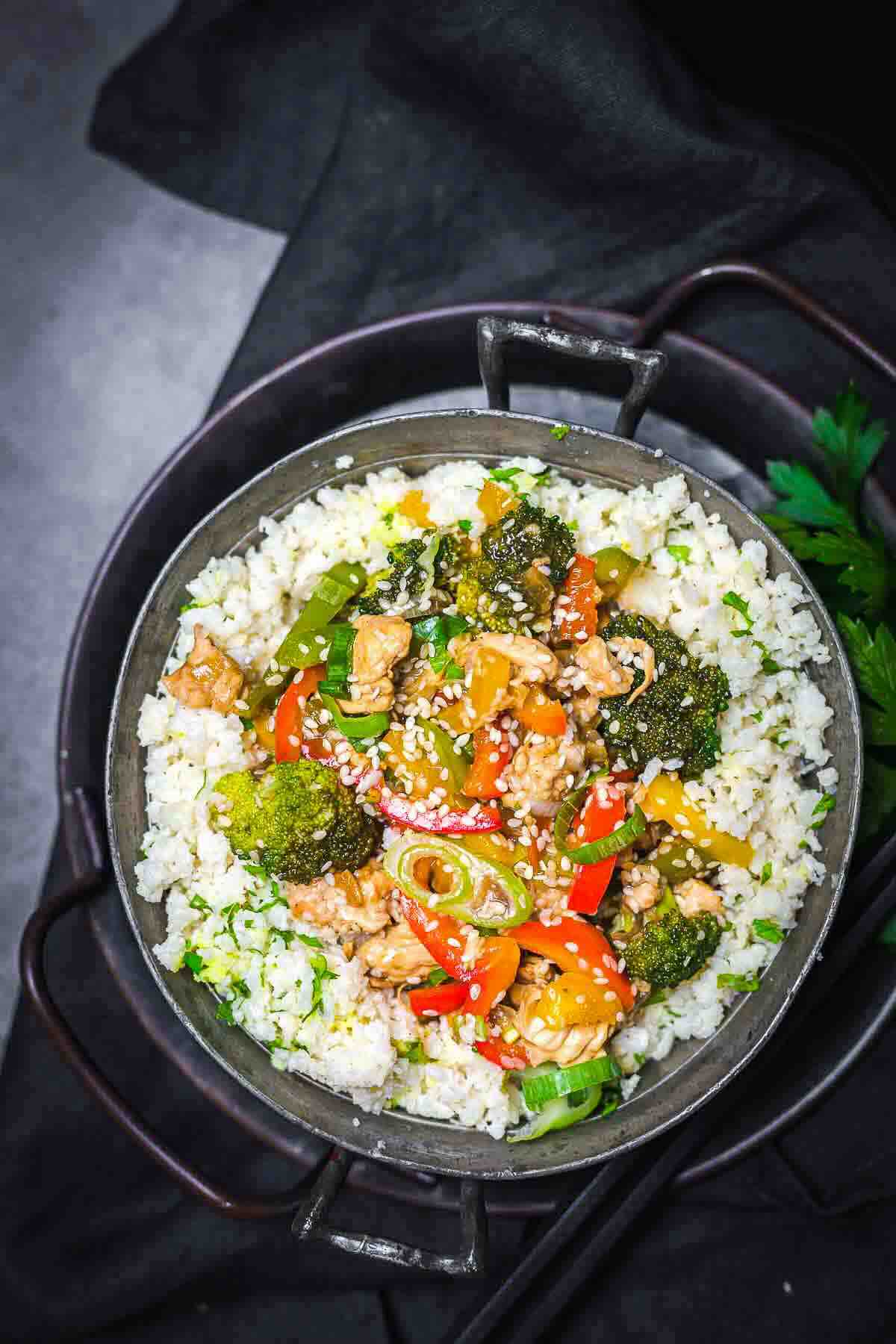 A bowl of stir-fried chicken, broccoli, bell peppers, and sesame seeds served over cauliflower rice.