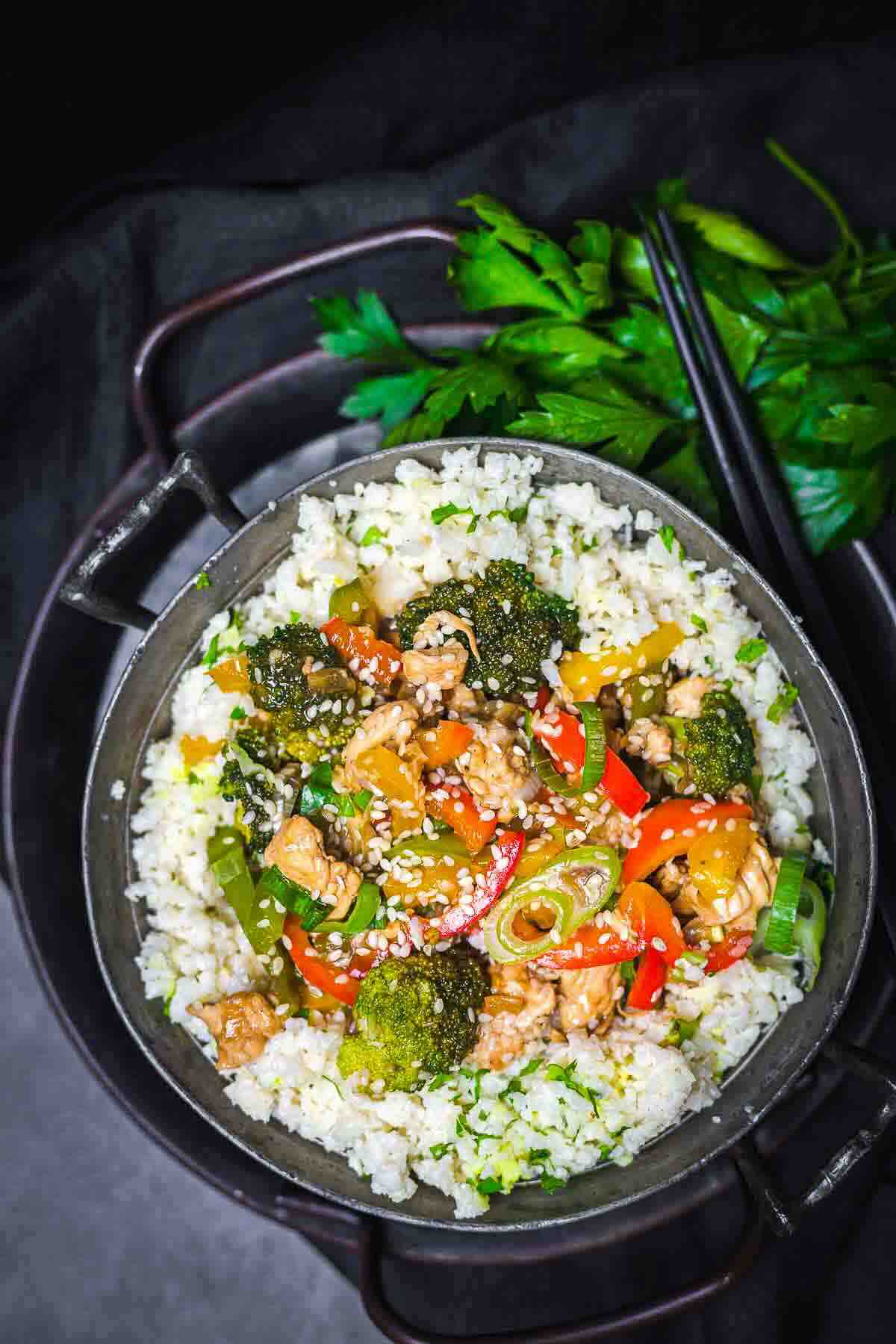 A bowl of stir-fried vegetables and chicken served over white rice, garnished with sesame seeds and spring onions, with fresh parsley and chopsticks beside the bowl.