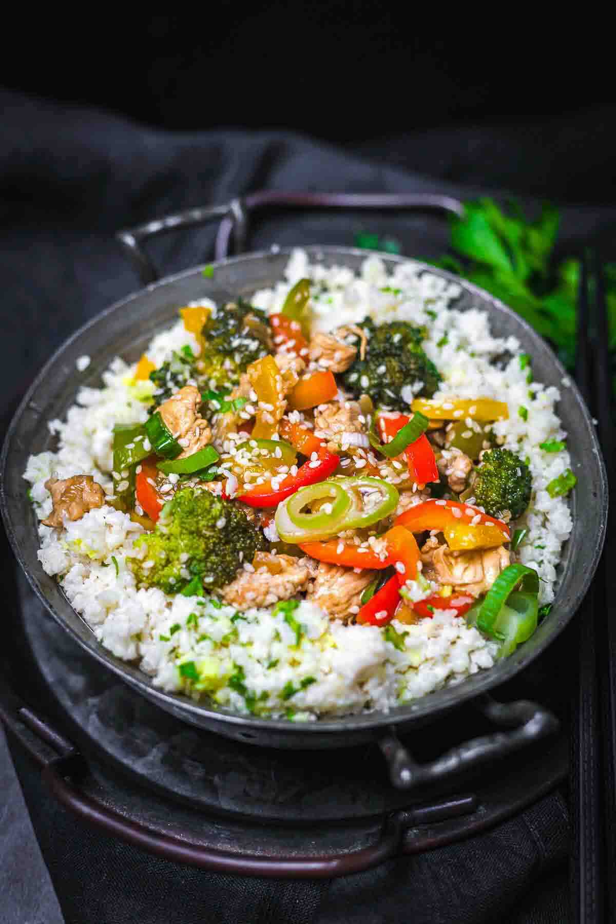 A bowl of stir-fried vegetables and chicken with sliced peppers, broccoli, and sesame seeds served over white rice, with chopsticks beside the bowl.