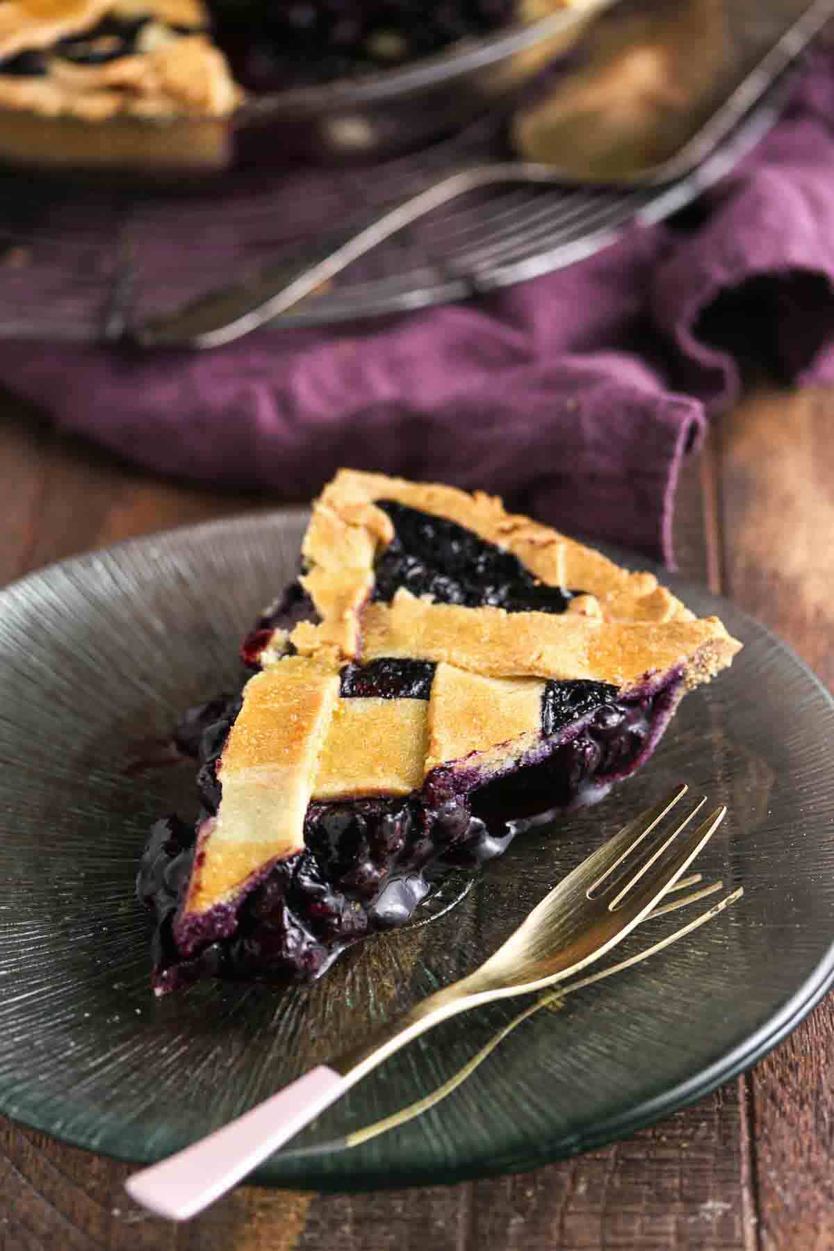 A slice of blueberry pie with a lattice crust on a glass plate, next to a gold fork, with a purple cloth in the background.