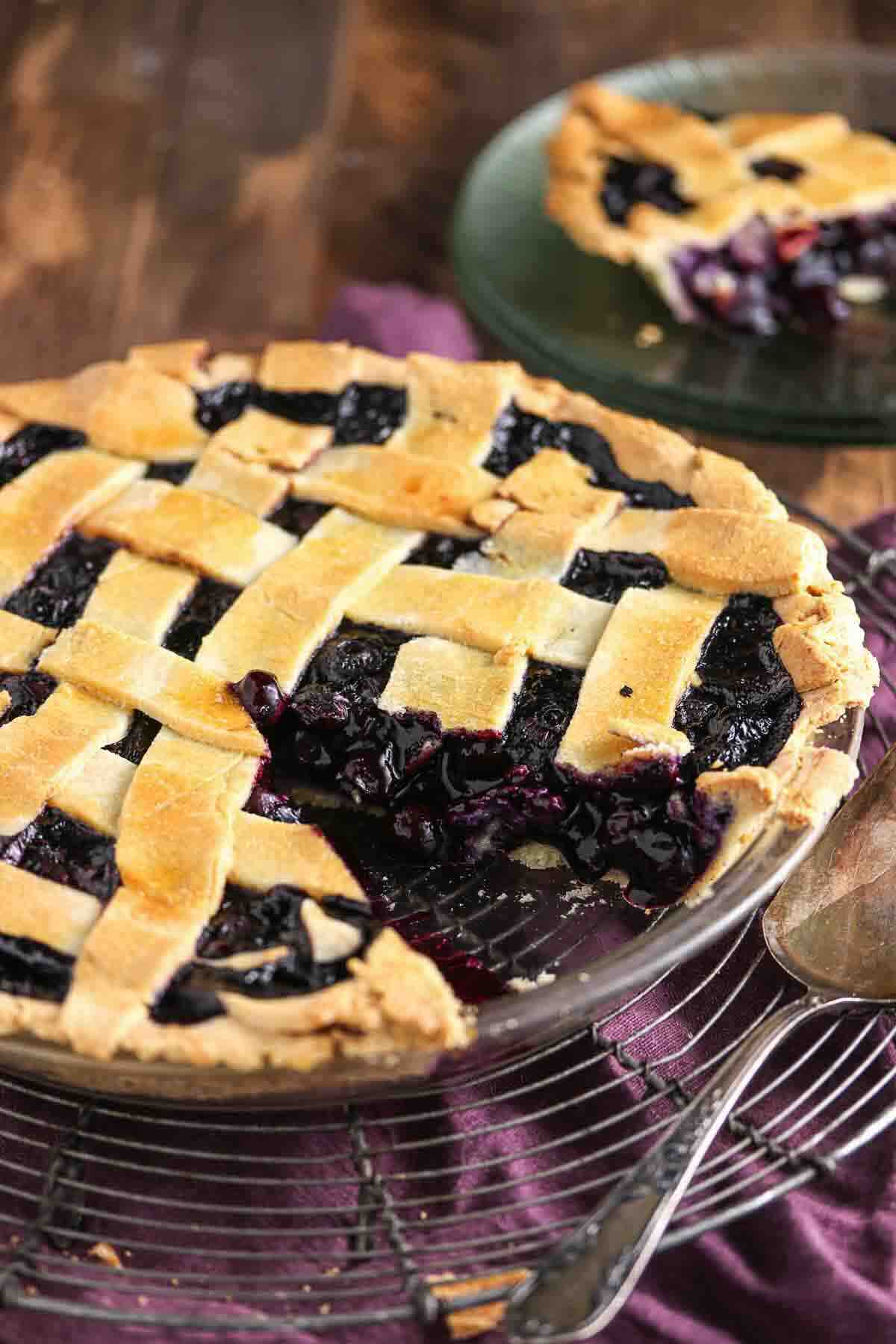 A lattice-topped blueberry pie sits on a cooling rack with a slice missing; the removed slice is on a glass plate in the background.