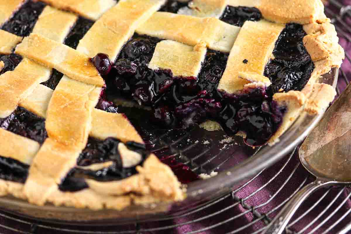 A blueberry pie with a lattice crust, one slice removed, sits on a wire cooling rack with a serving spoon nearby.