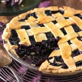 A lattice-topped blueberry pie in a glass dish with one slice removed, sitting on a wire rack with a serving utensil nearby.