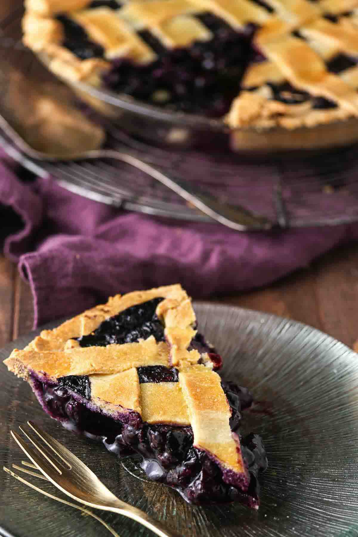 A slice of blueberry pie with a lattice crust is served on a plate with a fork, with the rest of the pie in the background.