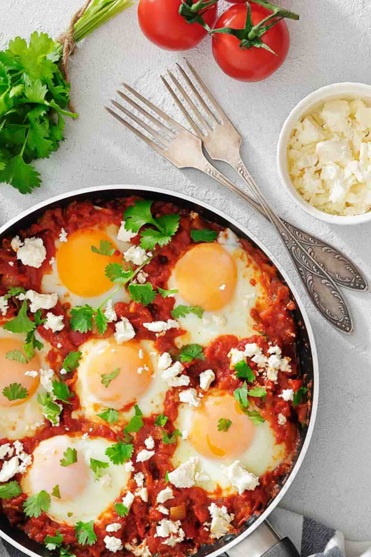 A pan of shakshuka with poached eggs in tomato sauce, garnished with cilantro and crumbled cheese. Fresh tomatoes, cilantro, and two forks are nearby on a light surface.