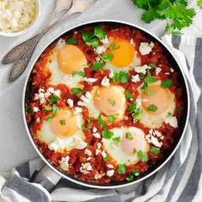 A skillet with six eggs cooked in a tomato sauce, topped with crumbled cheese and fresh herbs, sits on a striped towel next to utensils and a bowl of cheese.