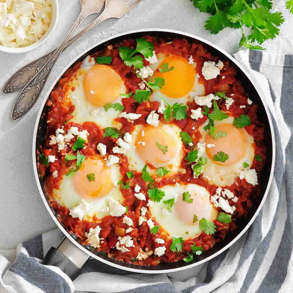A skillet with six eggs cooked in a tomato sauce, topped with crumbled cheese and fresh herbs, sits on a striped towel next to utensils and a bowl of cheese.