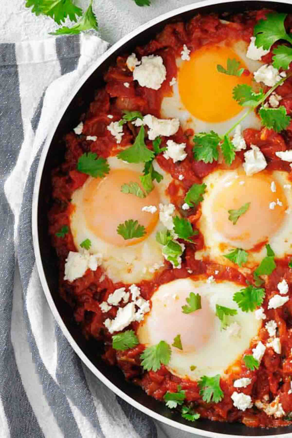 A skillet of shakshuka with four poached eggs in tomato sauce, topped with crumbled cheese and fresh cilantro, on a striped cloth.