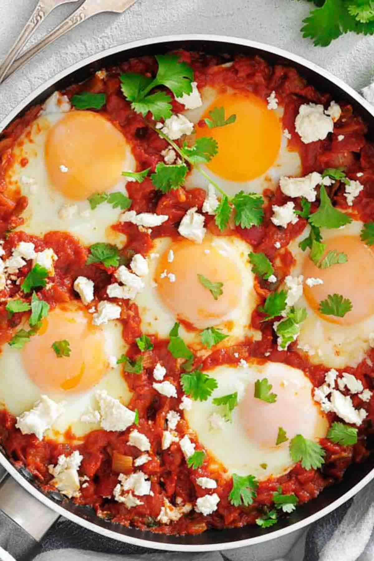 A skillet with six poached eggs in a tomato sauce, topped with cilantro and crumbled cheese.