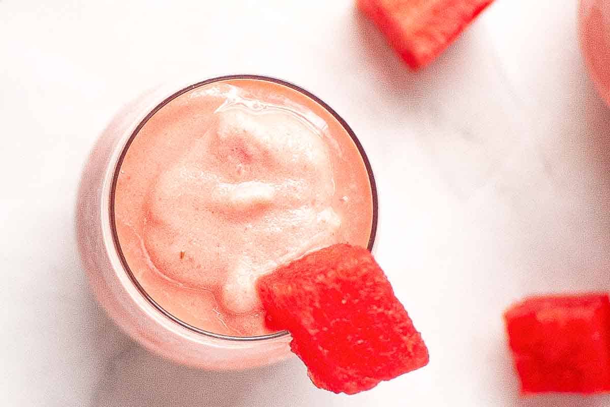 A glass of watermelon sherbet topped with a red watermelon chunk, placed on a white surface with additional watermelon pieces nearby.