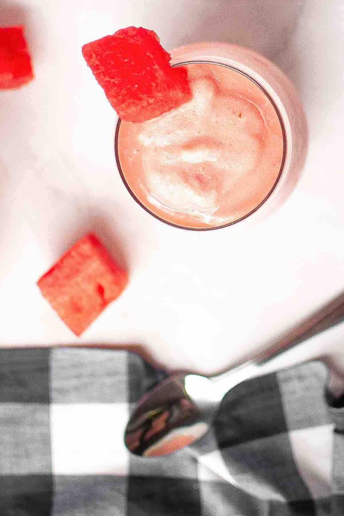 A glass of pink smoothie topped with a watermelon cube, with more watermelon pieces, a spoon, and a black-and-white checkered cloth nearby.
