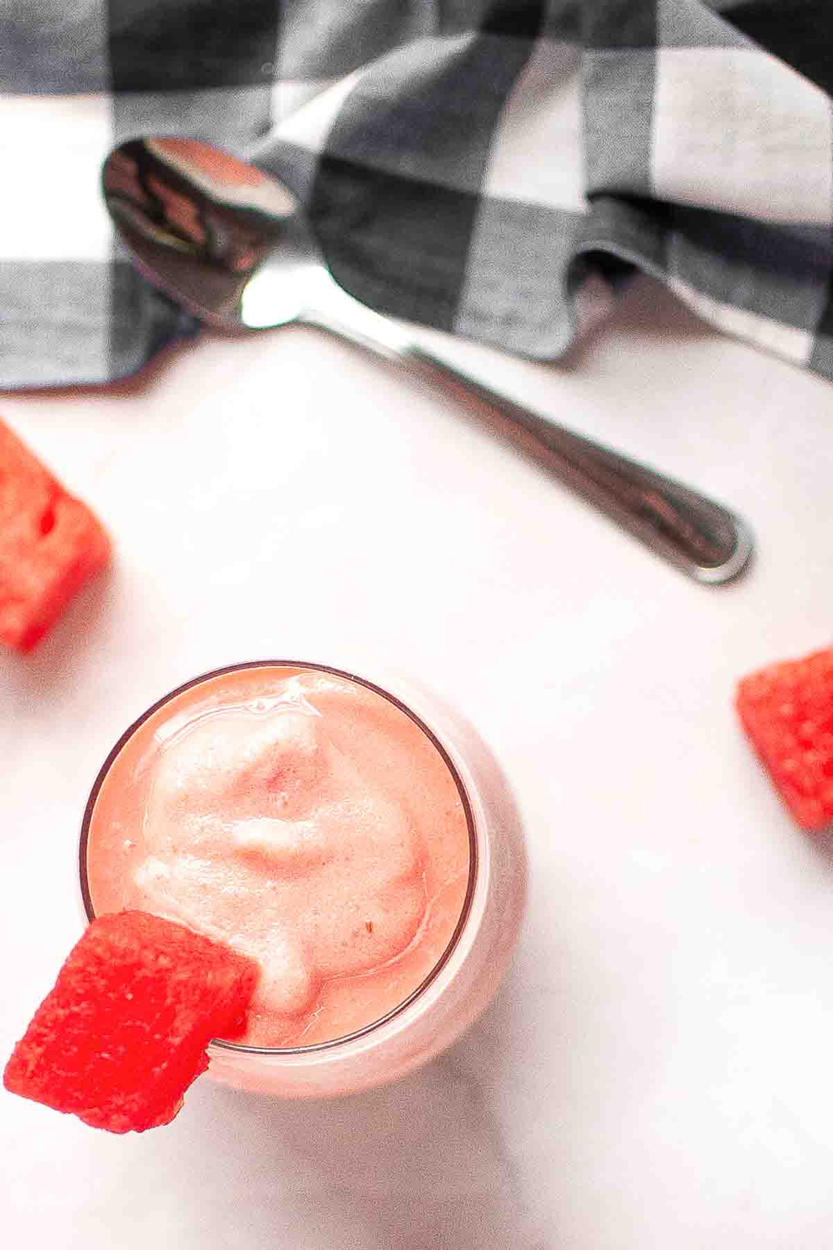 A glass of pink watermelon smoothie garnished with a watermelon chunk, with a spoon, checkered cloth, and watermelon pieces on a white surface.