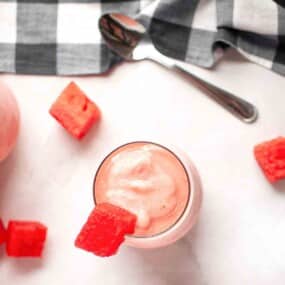 A glass of pink smoothie topped with a watermelon cube, surrounded by more watermelon cubes, a spoon, and a checkered cloth on a white surface.