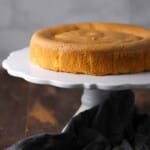 A plain, round sponge cake sits on a white cake stand with a dark cloth draped nearby on a wooden surface.