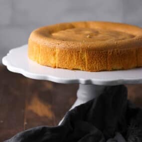 A plain, round sponge cake sits on a white cake stand with a dark cloth draped nearby on a wooden surface.