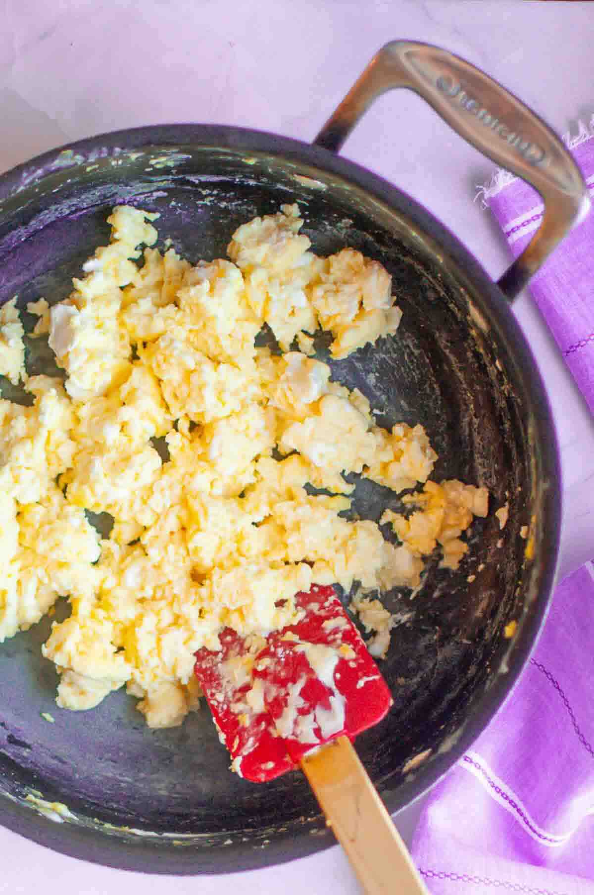 A pan of scrambled eggs with a red spatula, placed on a light surface next to a purple and white cloth.