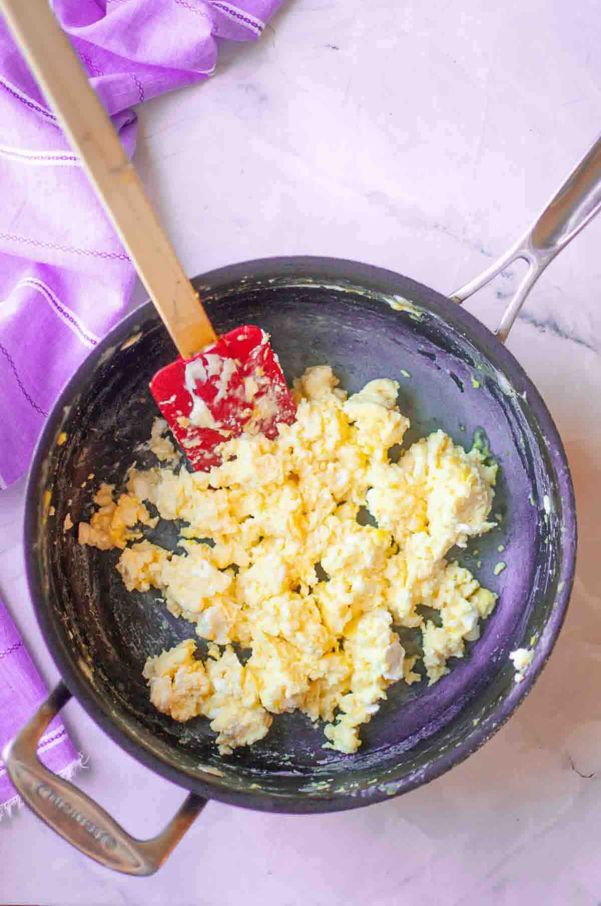 Scrambled eggs in a black pot with a red spatula, sitting on a light marble countertop next to a folded purple cloth.