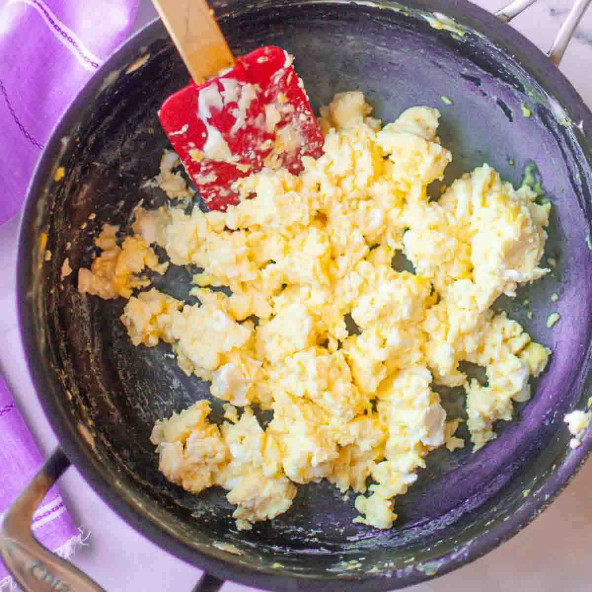 A pan filled with scrambled eggs being stirred with a red spatula, placed on a stove with a purple cloth nearby.