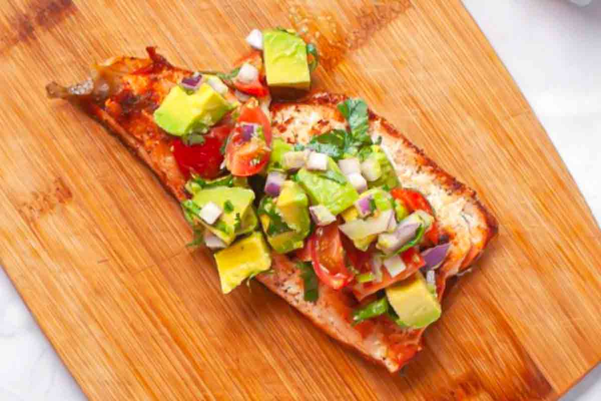 Grilled salmon fillet topped with a chunky salsa of avocado, cherry tomatoes, red onion, and herbs, served on a wooden cutting board.