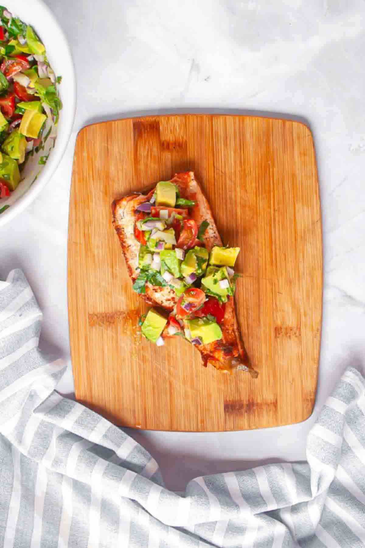 Grilled fish fillet topped with diced avocado, tomato, and herbs, served on a wooden board next to a gray and white striped cloth.