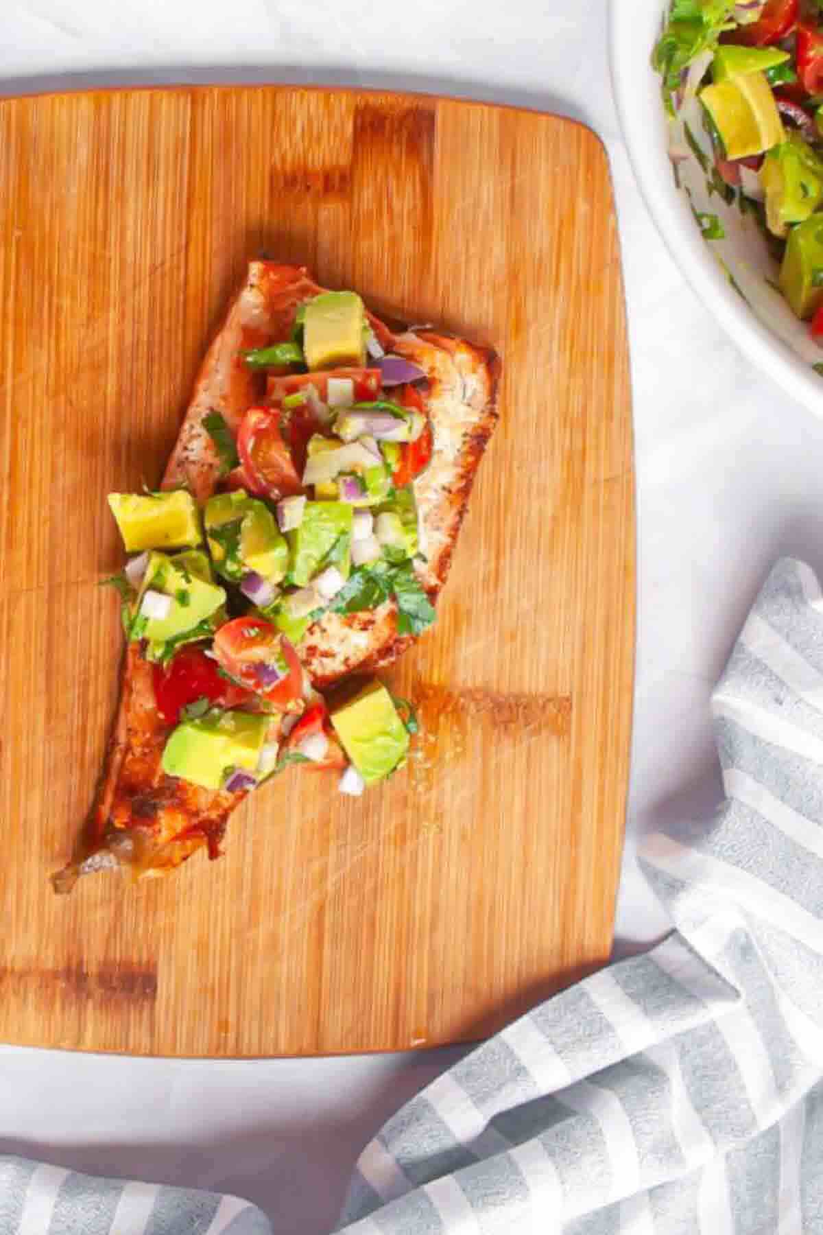 Grilled salmon fillet topped with diced avocado, tomatoes, onions, and herbs on a wooden cutting board, next to a bowl of salad and a striped cloth.
