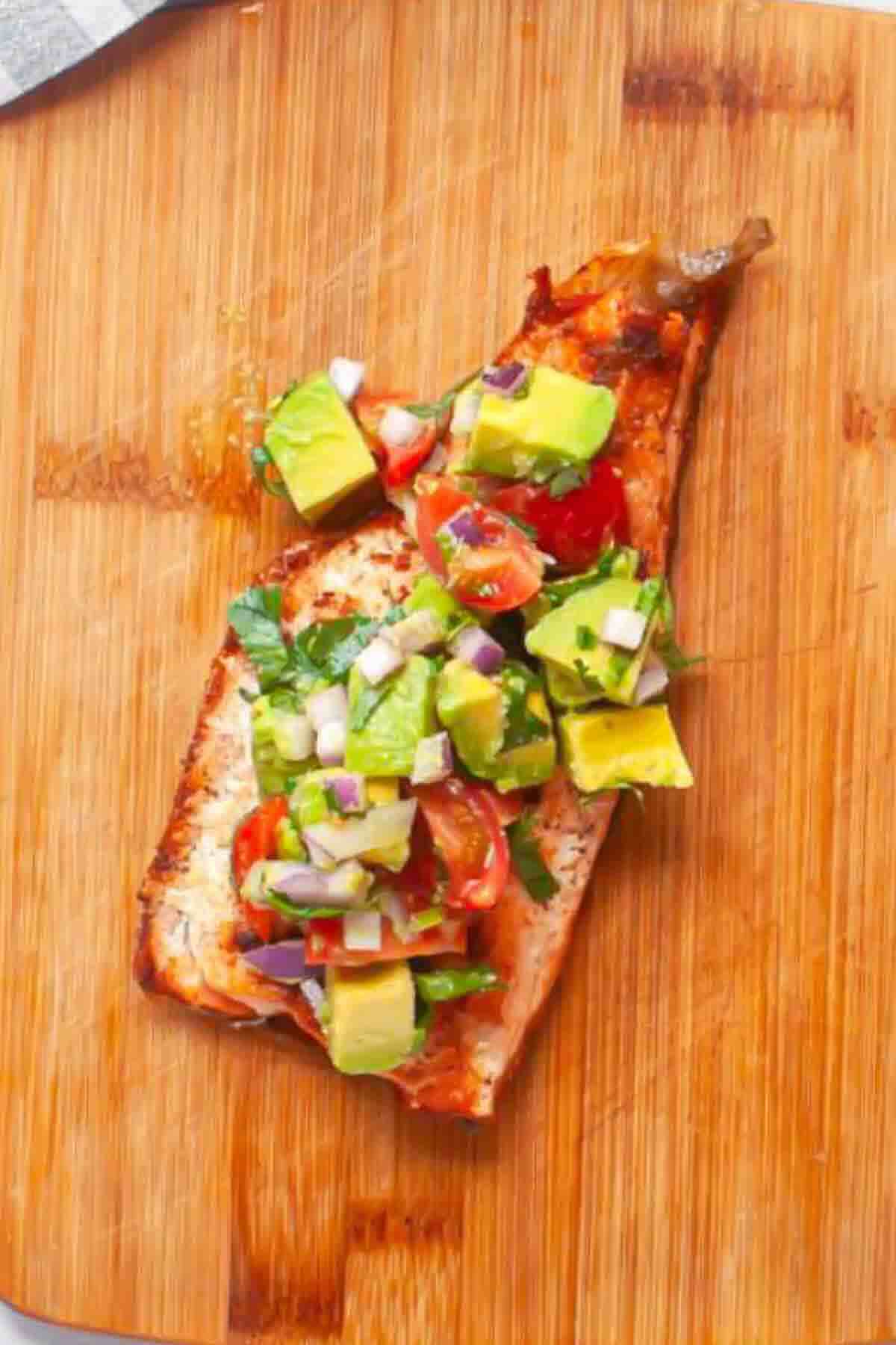 A cooked salmon fillet topped with diced avocado, tomato, red onion, and herbs sits on a wooden cutting board.