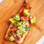 Grilled salmon fillet topped with diced avocado, cherry tomatoes, red onion, and herbs on a wooden cutting board.