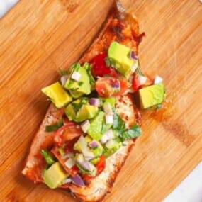 Grilled salmon fillet topped with diced avocado, cherry tomatoes, red onion, and herbs on a wooden cutting board.