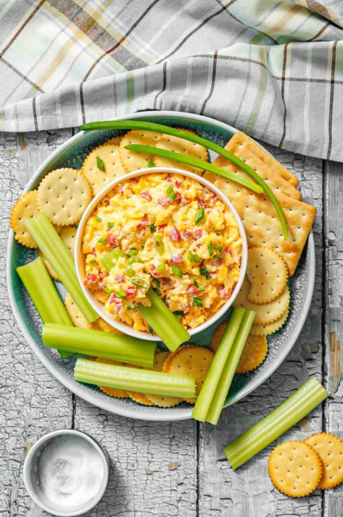 A bowl of pimento cheese spread surrounded by crackers and celery sticks on a plate, with a plaid cloth napkin in the background.