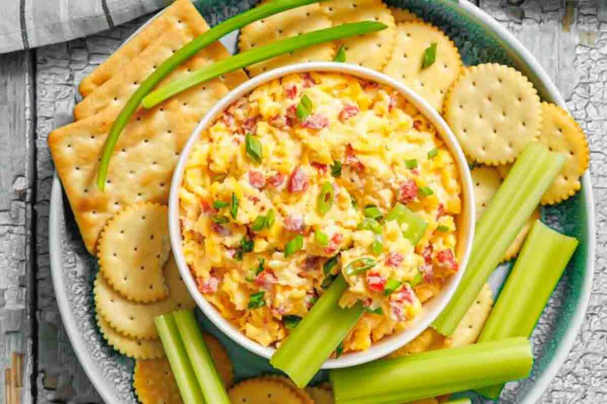 Bowl of pimento cheese dip garnished with green onions, surrounded by celery sticks and assorted round and square crackers on a blue plate.