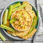 A bowl of pimento cheese dip is surrounded by celery sticks and round crackers on a plate, with a striped napkin beside it.