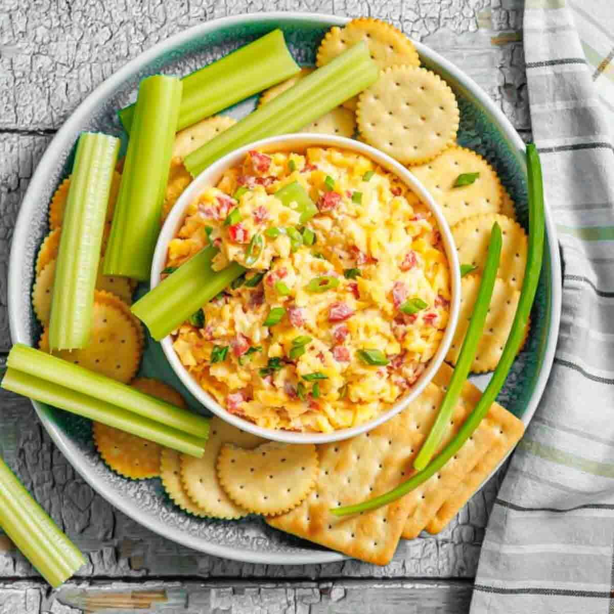 A bowl of pimento cheese dip is surrounded by celery sticks and round crackers on a plate, with a striped napkin beside it.
