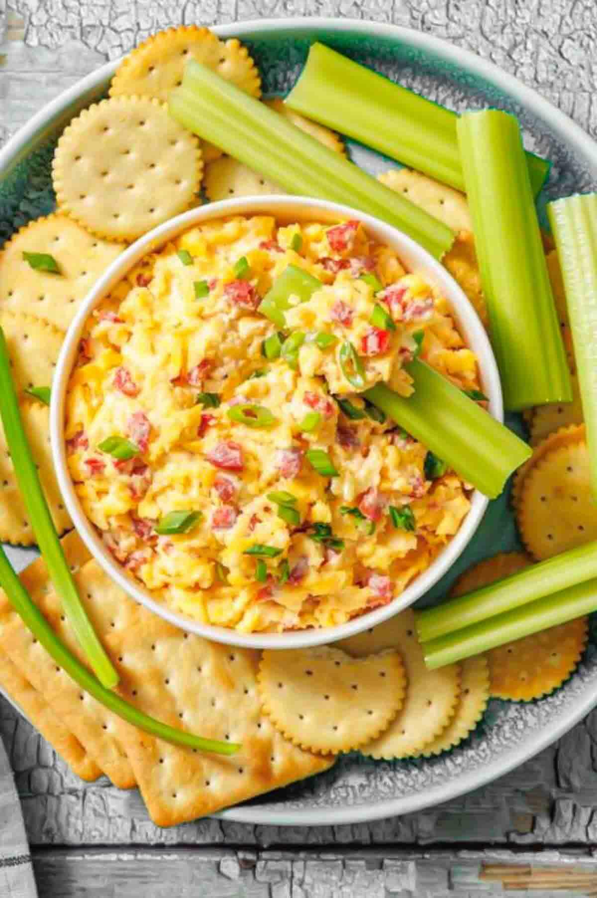 A bowl of pimento cheese spread is surrounded by round crackers, square crackers, and celery sticks on a plate.