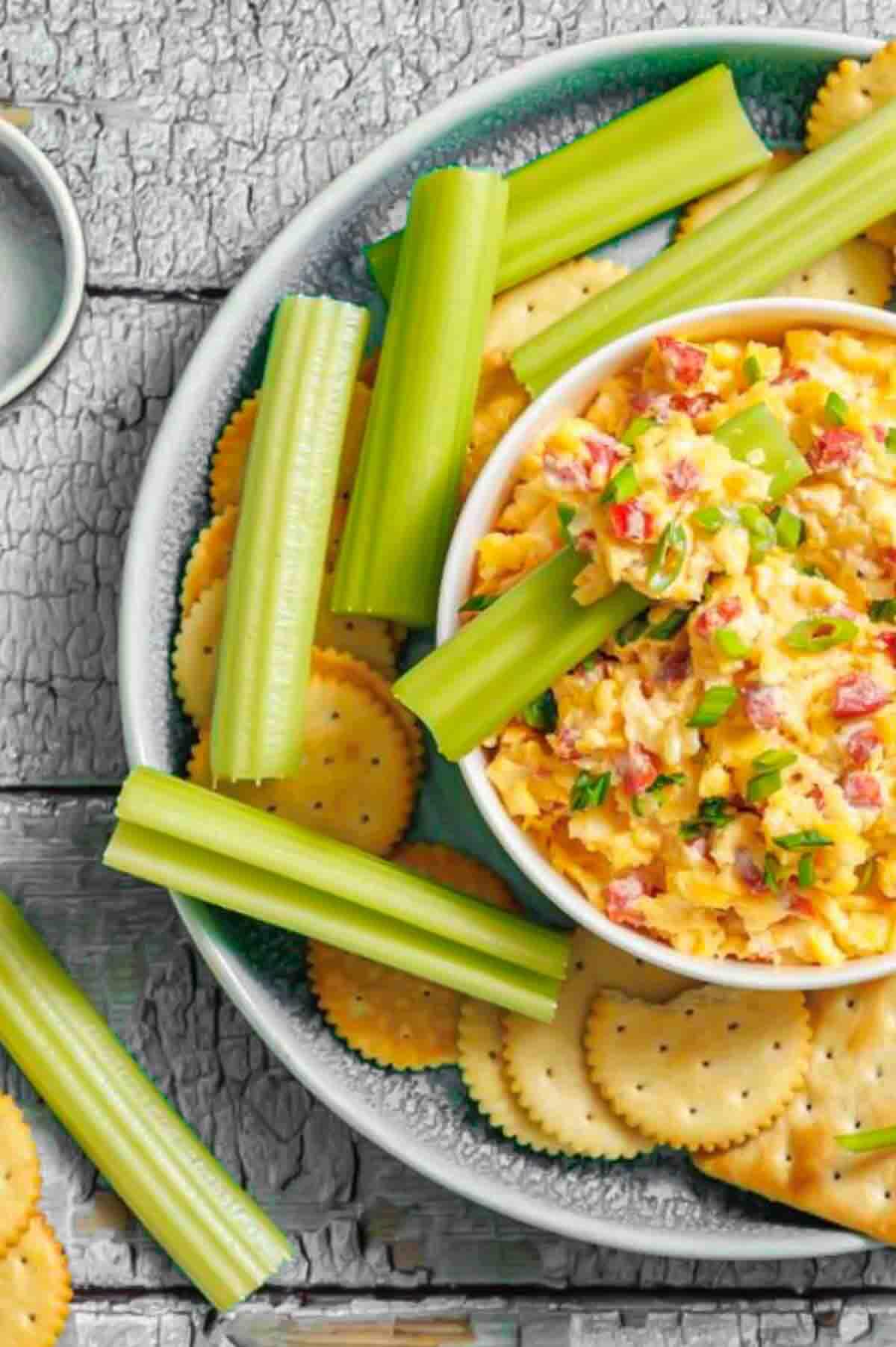 Bowl of pimento cheese dip surrounded by celery sticks and round crackers on a white plate.