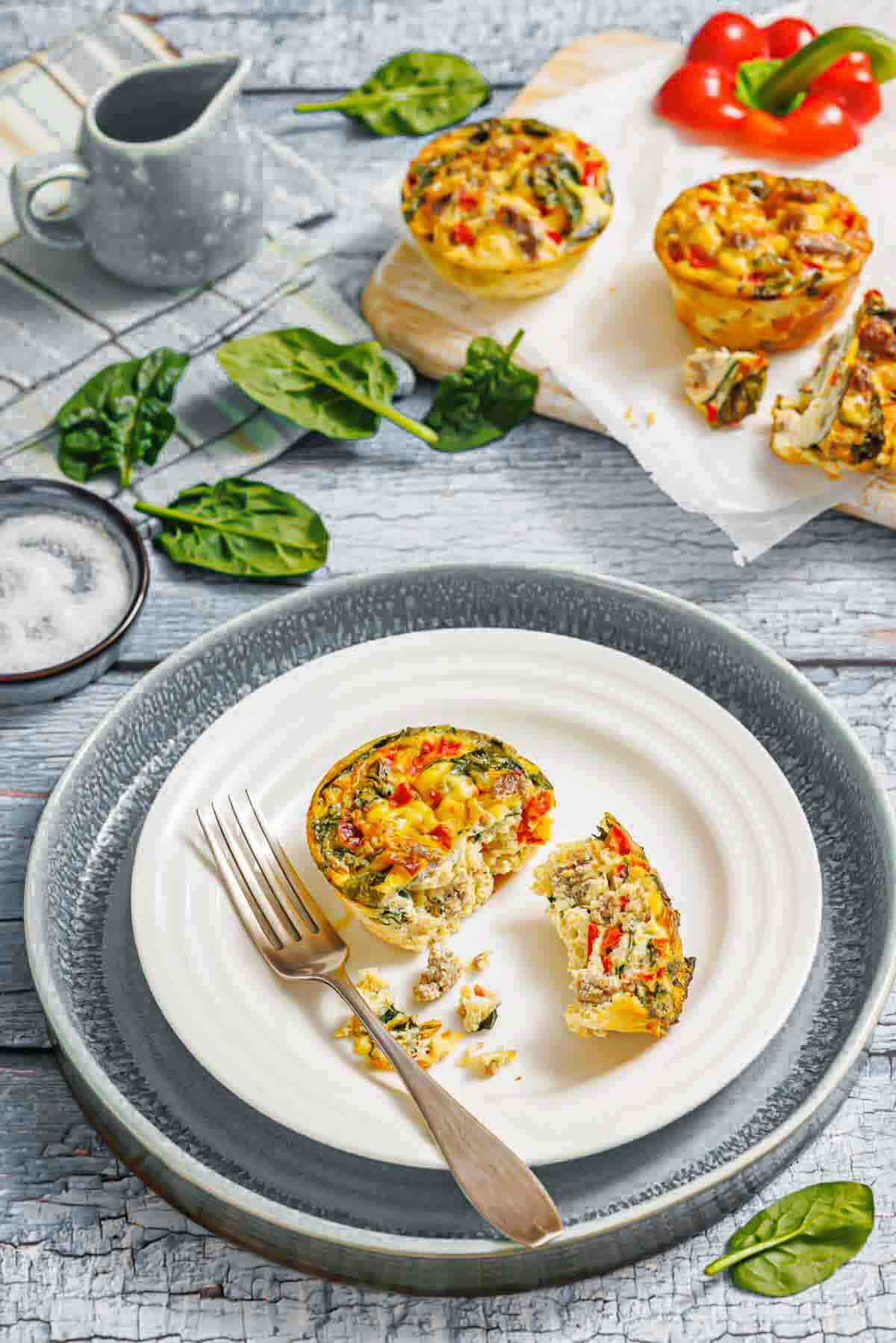 Mini vegetable frittata muffin on a plate with a fork, surrounded by fresh spinach leaves and more frittatas on a board in the background.